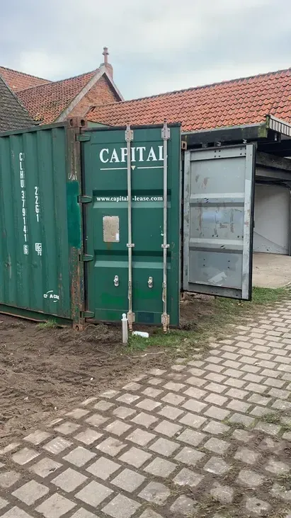 Grüner Schiffscontainer mit offener Tür neben einem Gebäude, beide im Freien.