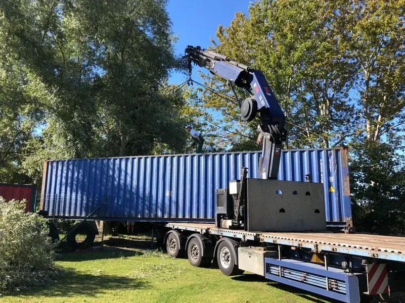 Ein blauer Schiffscontainer wird in einer grasbewachsenen, von Bäumen umgebenen Fläche gehoben.