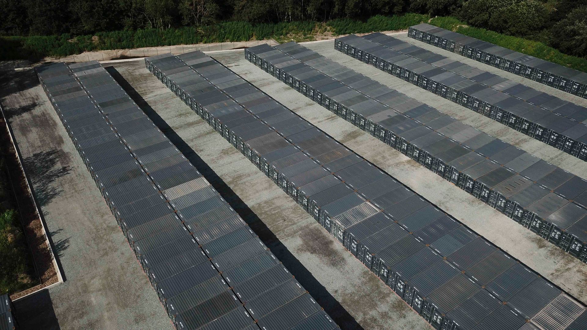Luftaufnahme einer Lagerhalle mit Reihen grauer Metallcontainer auf einer Betonfläche, umgeben von Bäumen.