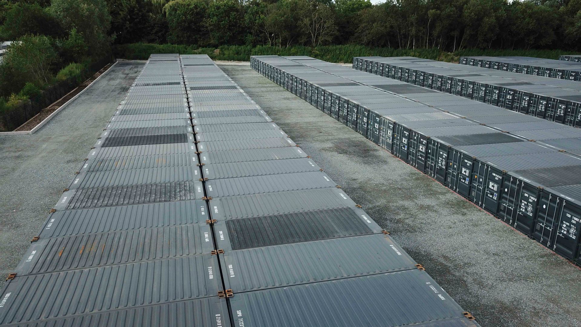 Reihen dunkelgrauer Schiffscontainer auf einer Betonfläche, im Hintergrund Bäume.