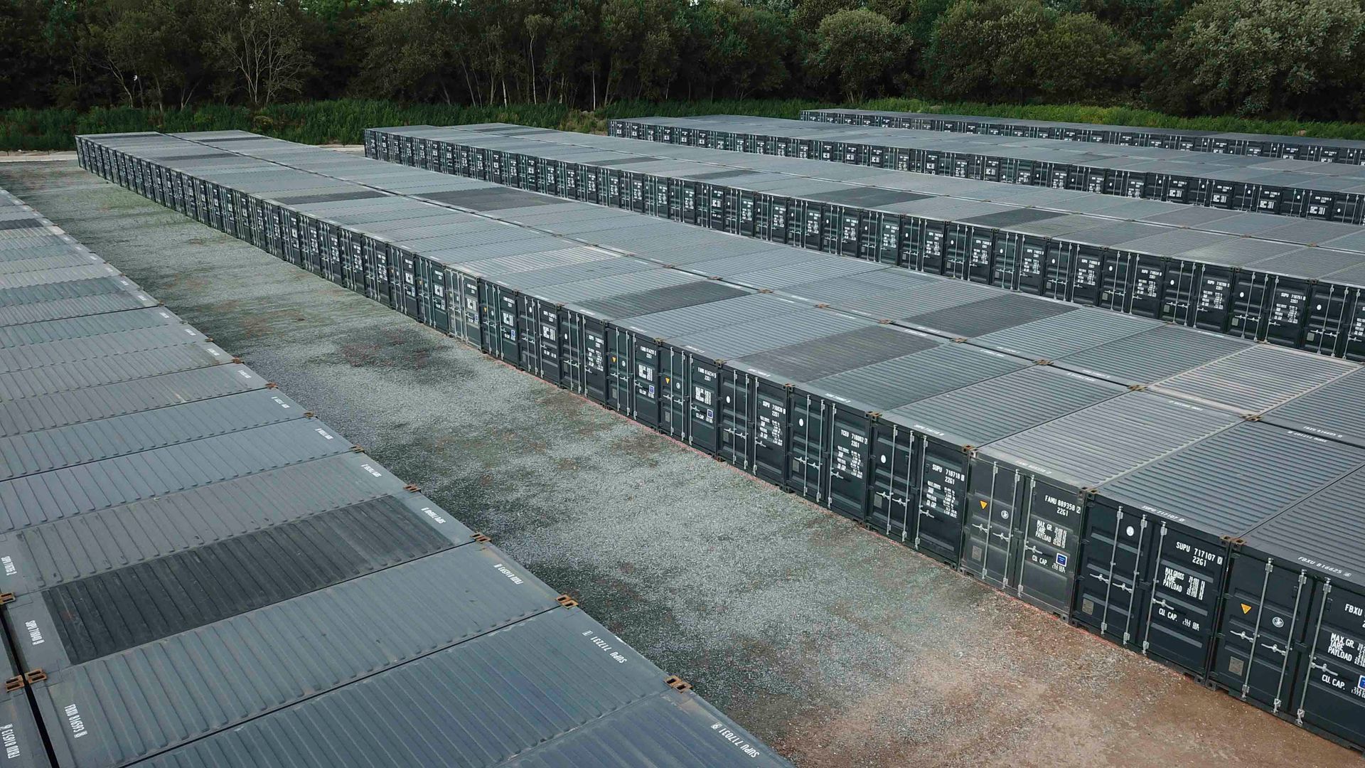 Reihen dunkler Schiffscontainer auf einer Kiesfläche, vermutlich ein Lagerplatz, im Hintergrund Bäume.