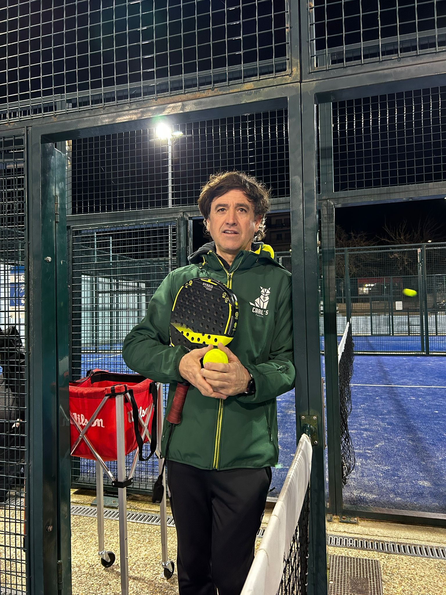 Un hombre con una chaqueta verde sostiene una raqueta de tenis y una pelota de tenis.
