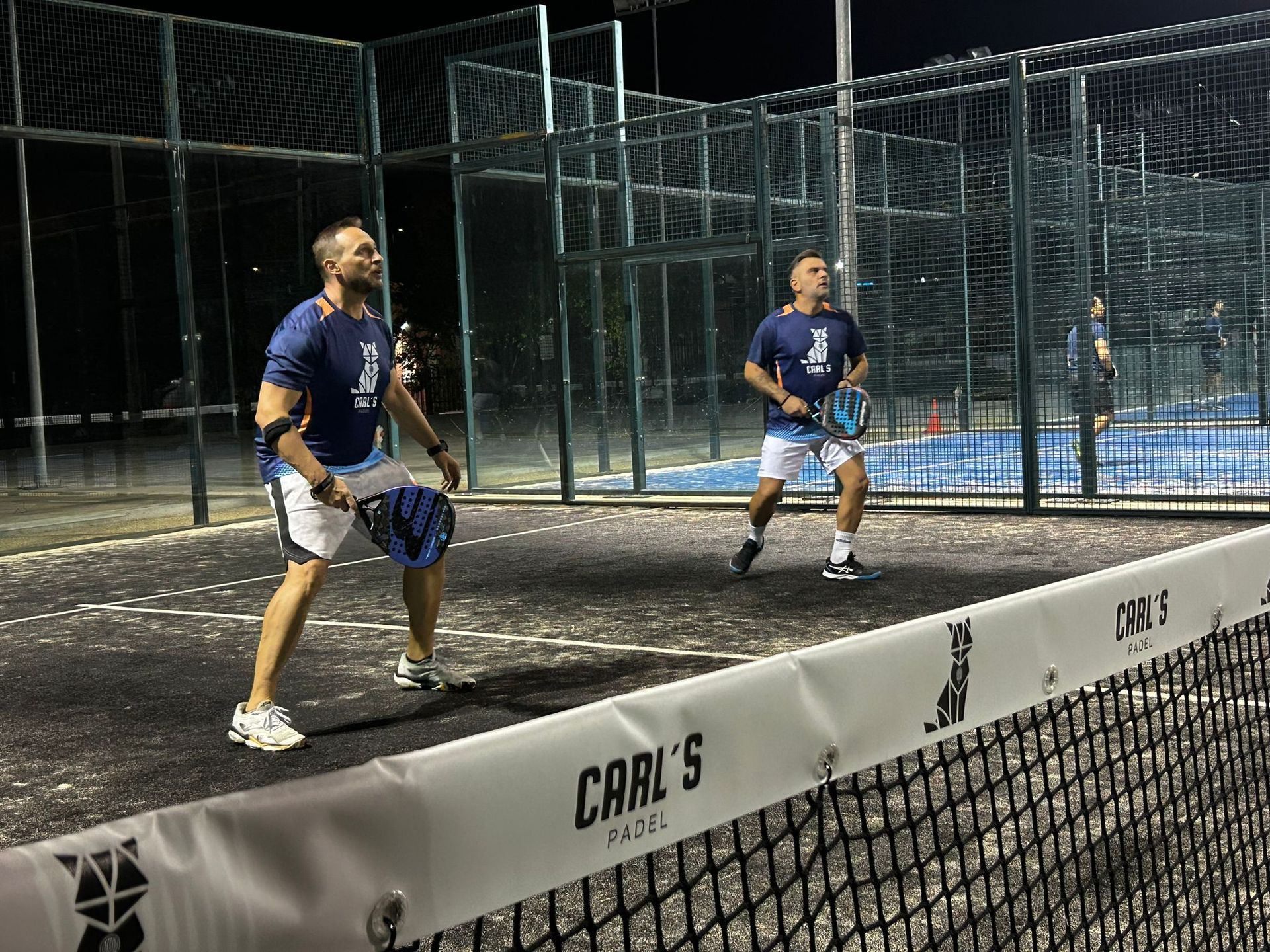Dos hombres están jugando al pádel en una cancha por la noche.