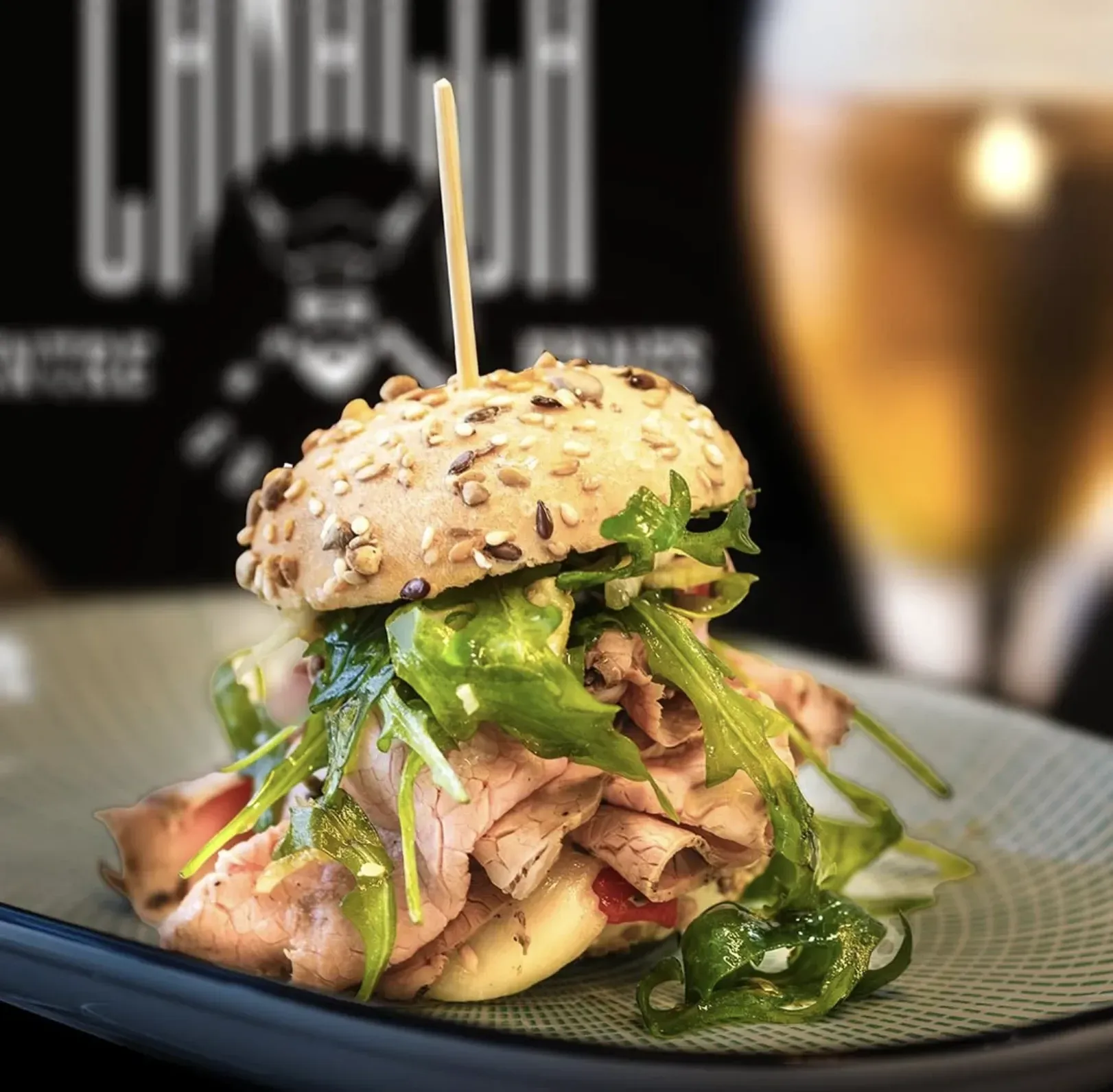 Un primer plano de un sándwich en un plato con un vaso de cerveza al fondo.