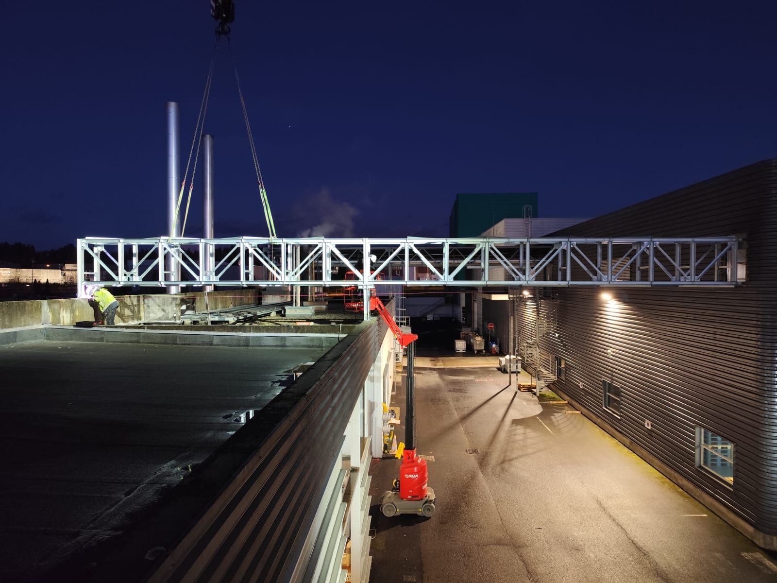 Installation nocturne d'une passerelle piétonne. Une grue soulève la structure métallique blanche au-dessus d'un bâtiment.