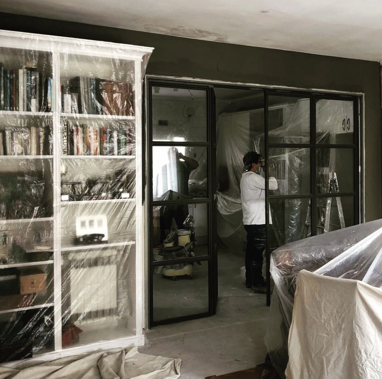 Habitación en reforma, estantería y puerta tapadas con plástico, dos personas trabajando, paredes oscuras.