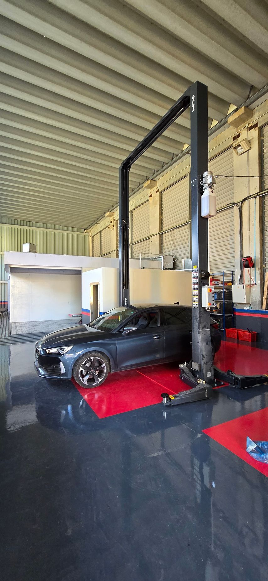Une voiture sombre sur une plateforme rouge dans un garage, près d'un monte-voiture. Le sol est sombre.