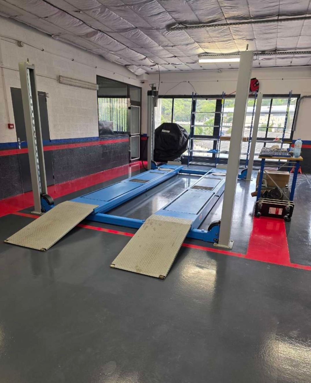 Un pont élévateur dans un garage avec rampes, sur un sol gris. Garnitures rouges, noires et bleues ; une fenêtre en arrière-plan.