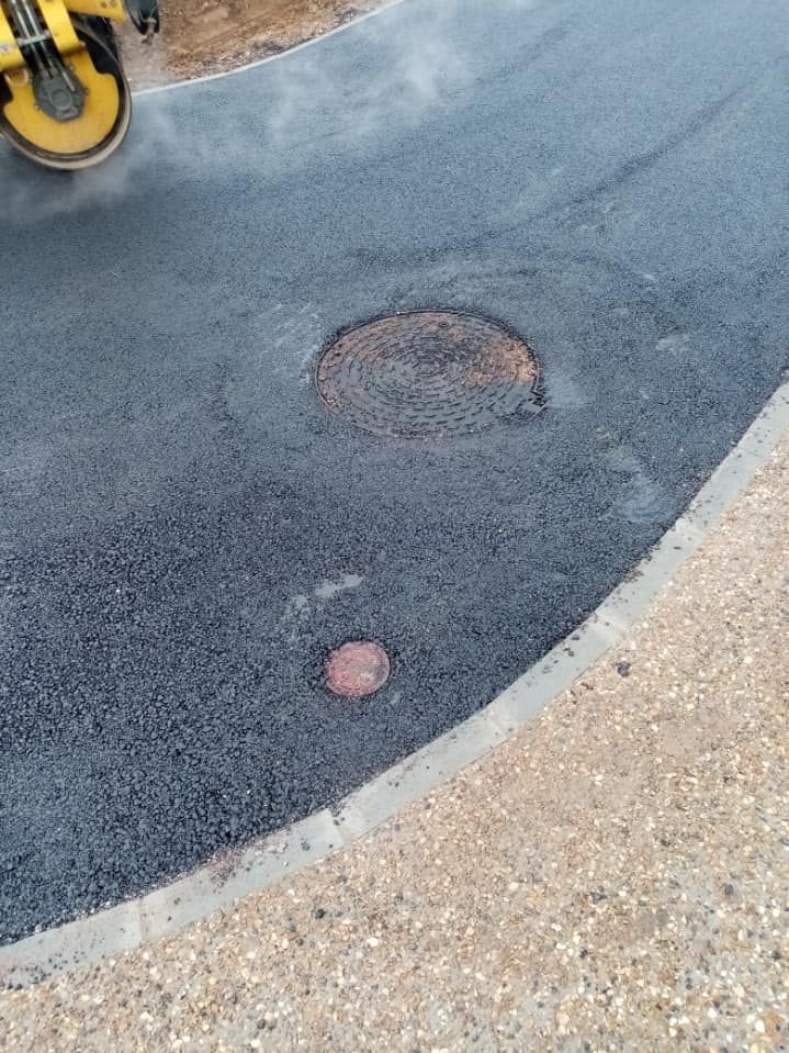 Une plaque d'égout et une petite vanne utilitaire circulaire dans de l'asphalte fraîchement posé, près d'une allée piétonne pavée de couleur claire.