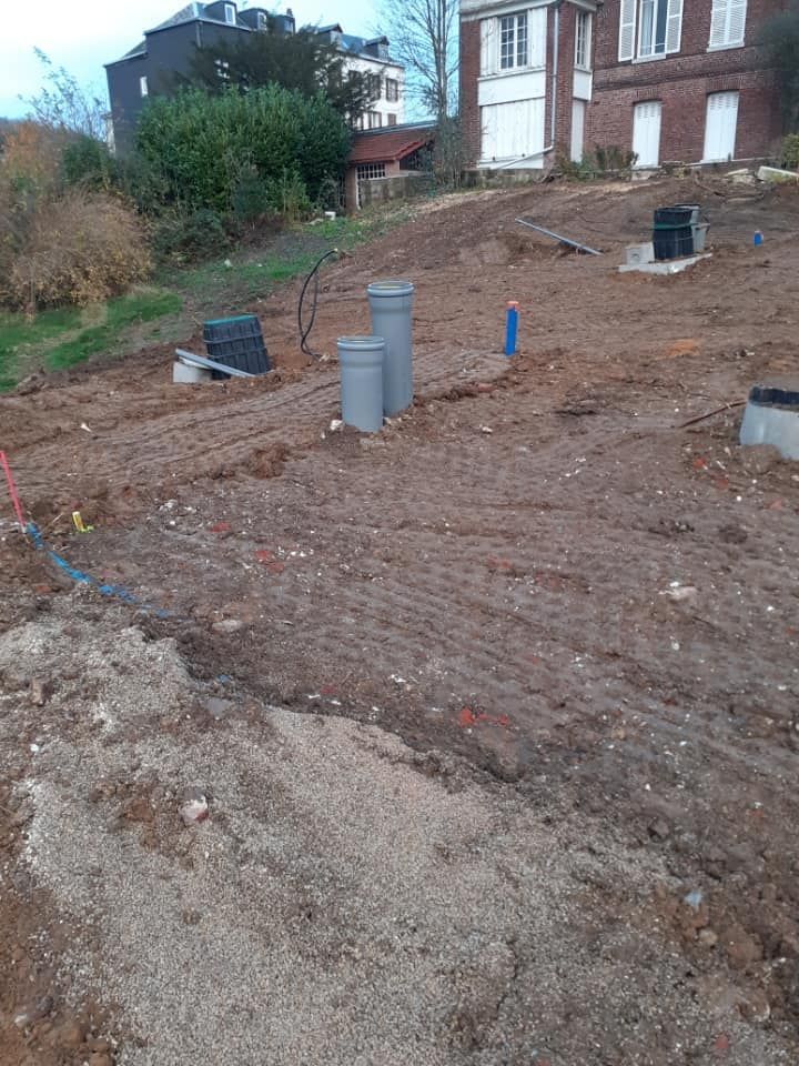 Un chantier en pente avec de la terre à nu, plusieurs tuyaux de drainage gris et une maison en briques en arrière-plan.