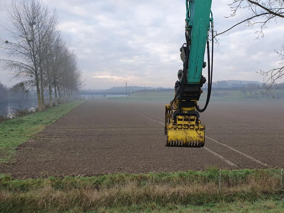 Par une journée brumeuse, un bras d'excavatrice turquoise muni d'une griffe mécanique jaune surplombe un champ labouré près d'une rangée d'arbres.