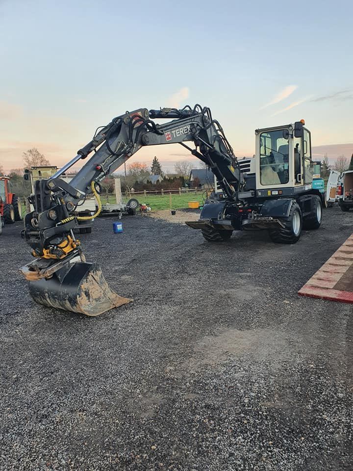 Une pelle mécanique grise sur roues, équipée d'un godet, est stationnée sur un terrain gravillonné en plein jour.