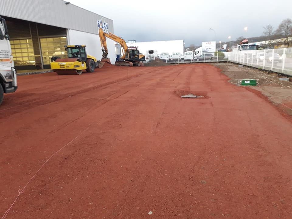 Un chantier de construction avec une surface de gravier rouge, un rouleau compresseur jaune et une excavatrice près d'un entrepôt.