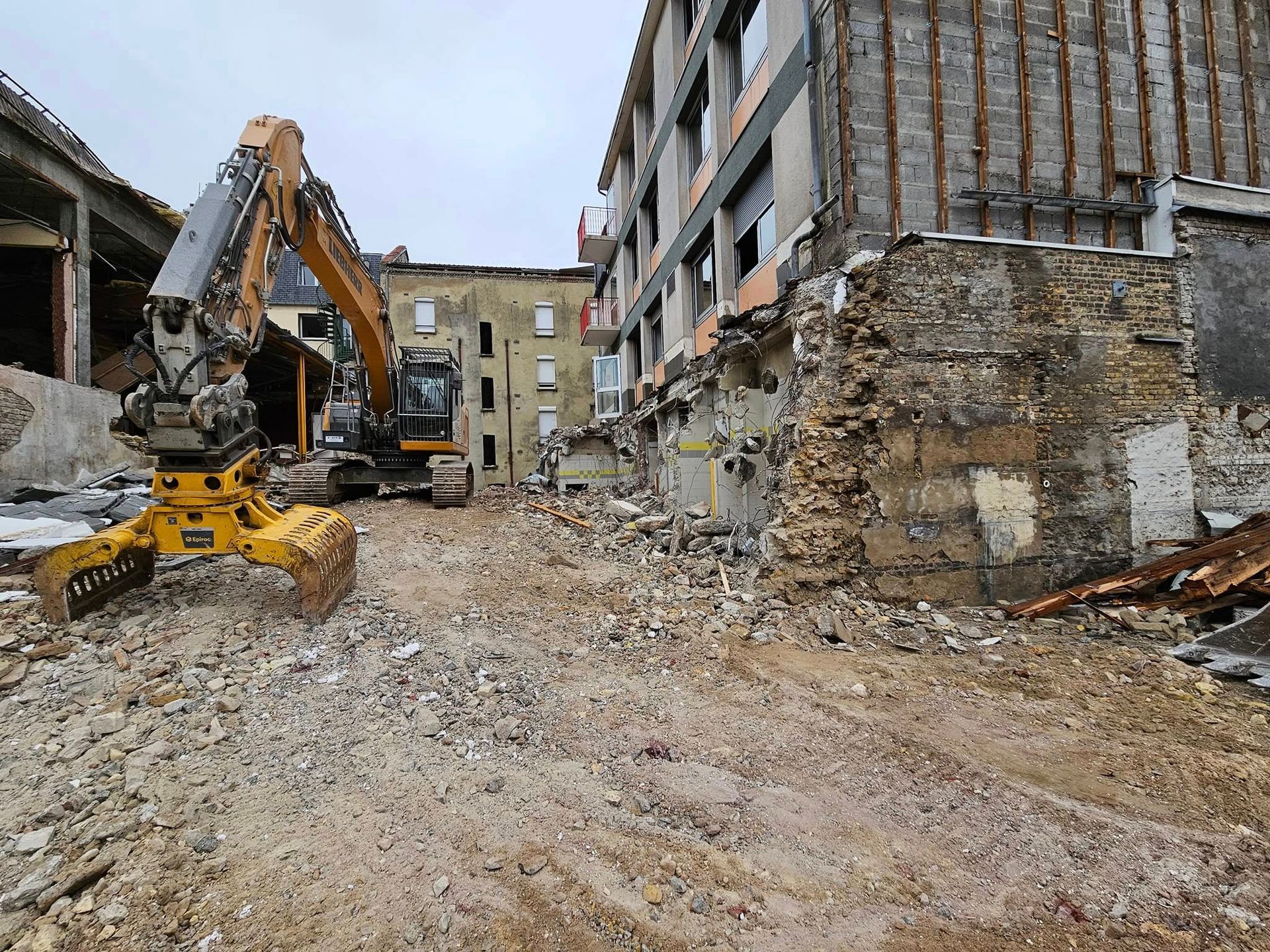Une pelleteuse jaune se trouve sur un chantier, au milieu des décombres, positionnée entre deux bâtiments partiellement démolis.