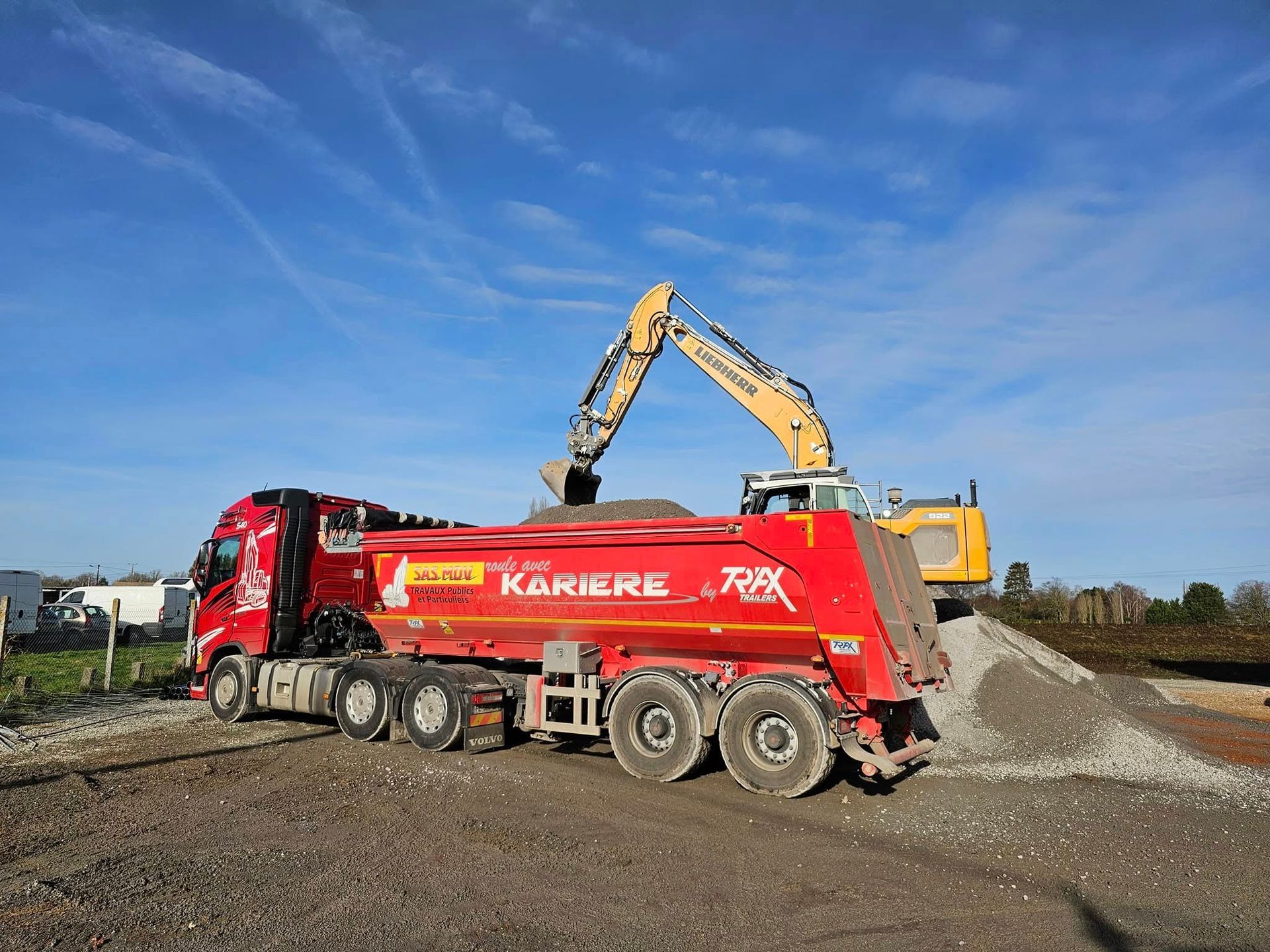 Une pelleteuse jaune charge du gravier dans la benne rouge d'un gros camion.