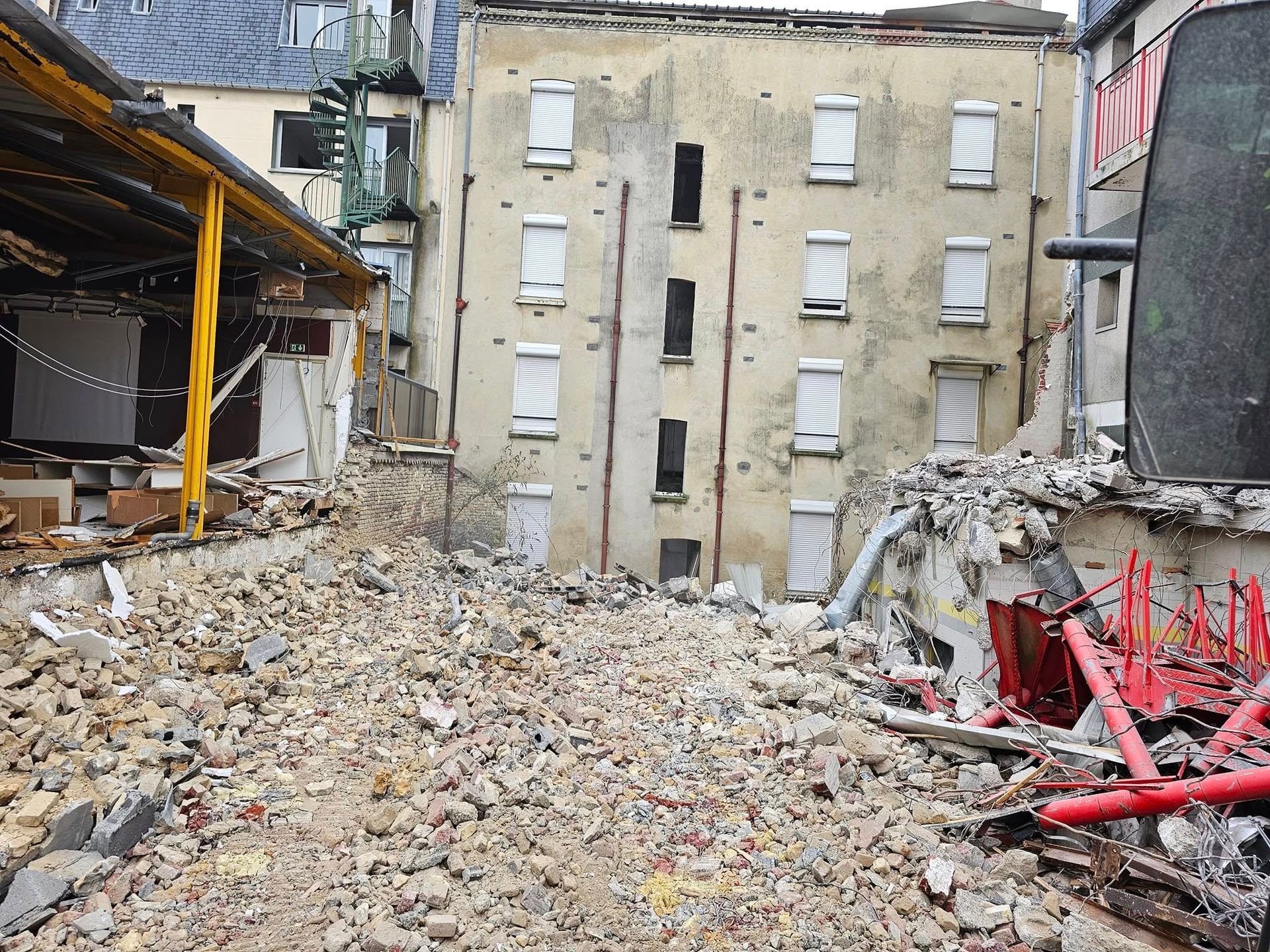 Débris et gravats devant un immeuble d'appartements beige aux fenêtres fermées par des volets blancs.
