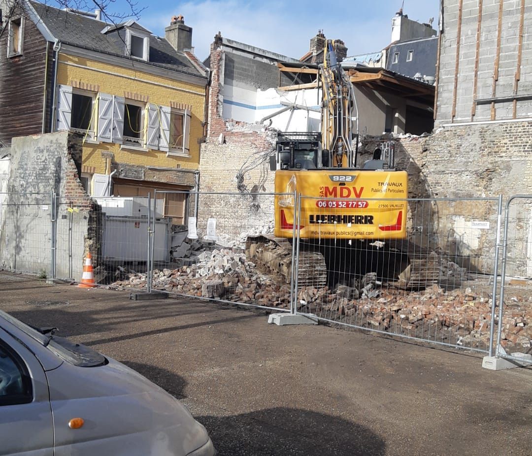 Une pelleteuse Liebherr jaune démolit un bâtiment jouxtant une maison existante, séparée par une clôture métallique temporaire.