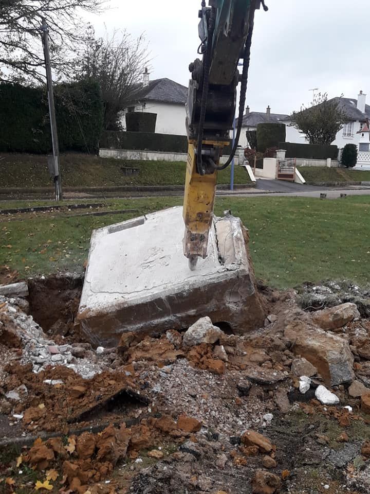 Un marteau hydraulique monté sur une excavatrice brise une grande dalle de béton dans un espace vert extérieur, en zone suburbaine.