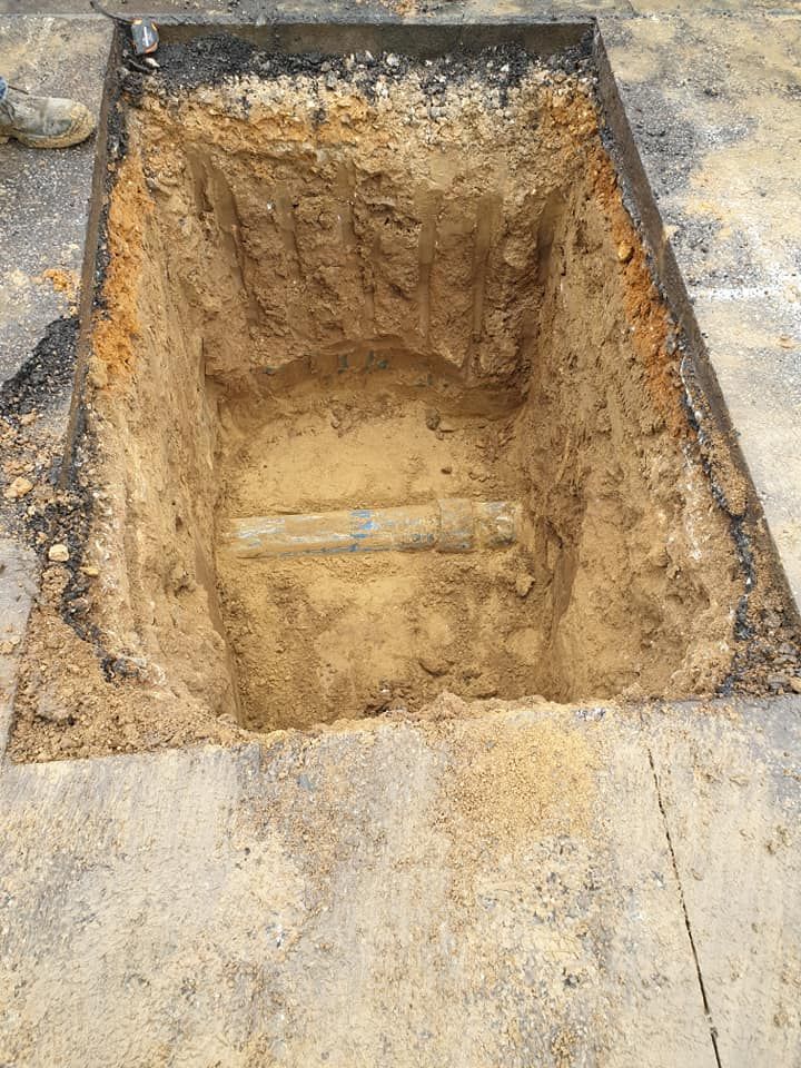 Une excavation rectangulaire pour services publics dans une surface pavée, mettant au jour un tuyau enfoui dans le sol sableux au fond.