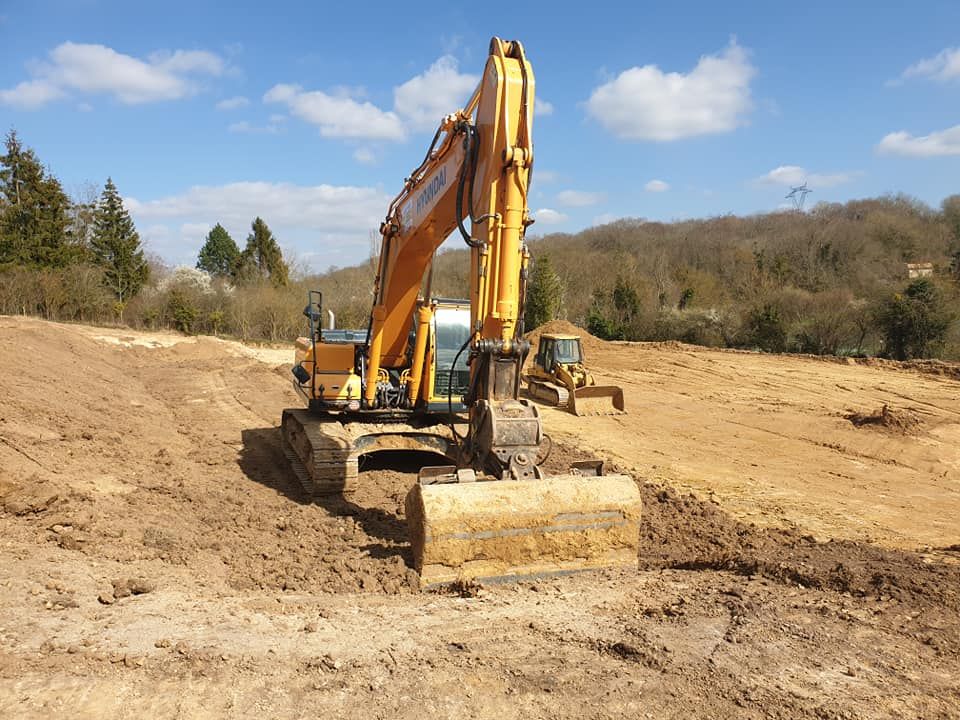 Une pelleteuse jaune à l'œuvre sur un grand chantier de construction ensoleillé, en terre battue, avec des collines et des arbres en arrière-plan.