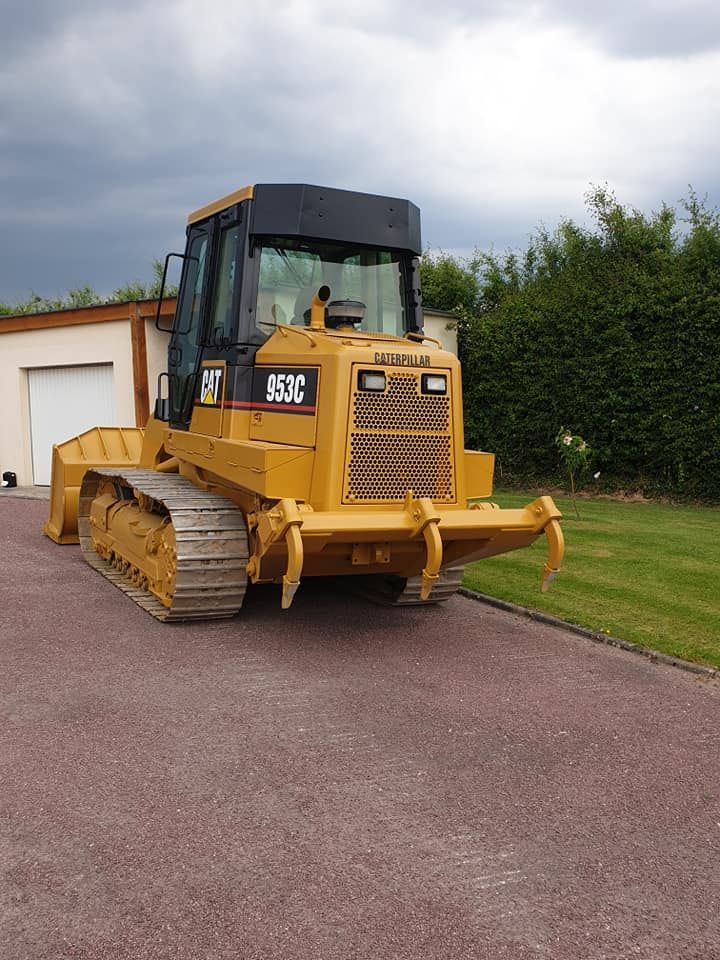 Une chargeuse sur chenilles jaune est garée sur une allée de gravier devant un bâtiment et de la verdure.