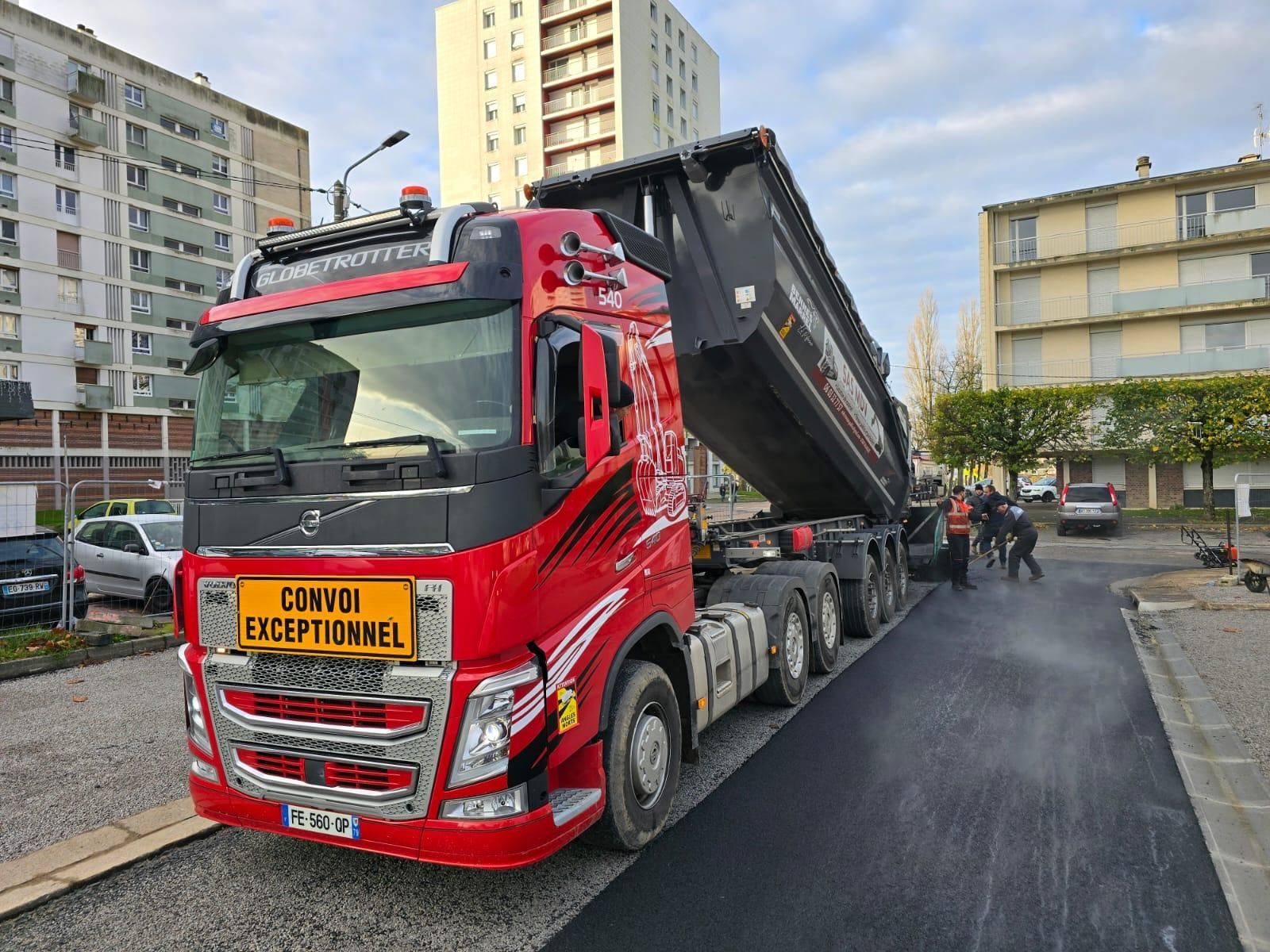 Un camion-benne Volvo rouge vif, sa remorque levée, est stationné sur un chantier de construction, entouré d'immeubles d'habitation.