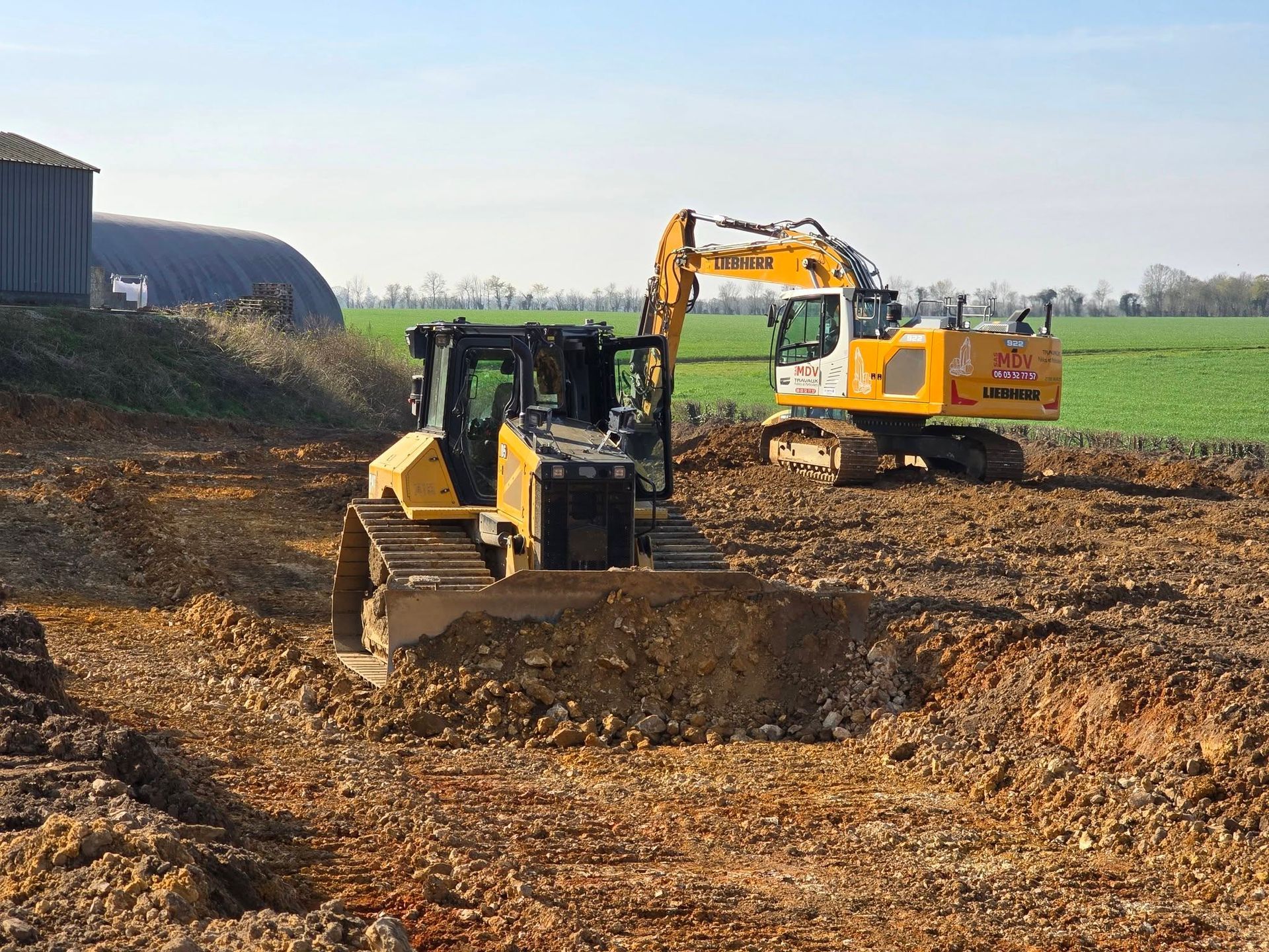 Un bulldozer et une excavatrice jaunes travaillent sur un chantier de construction en terre brune et accidentée, avec un champ vert en arrière-plan.