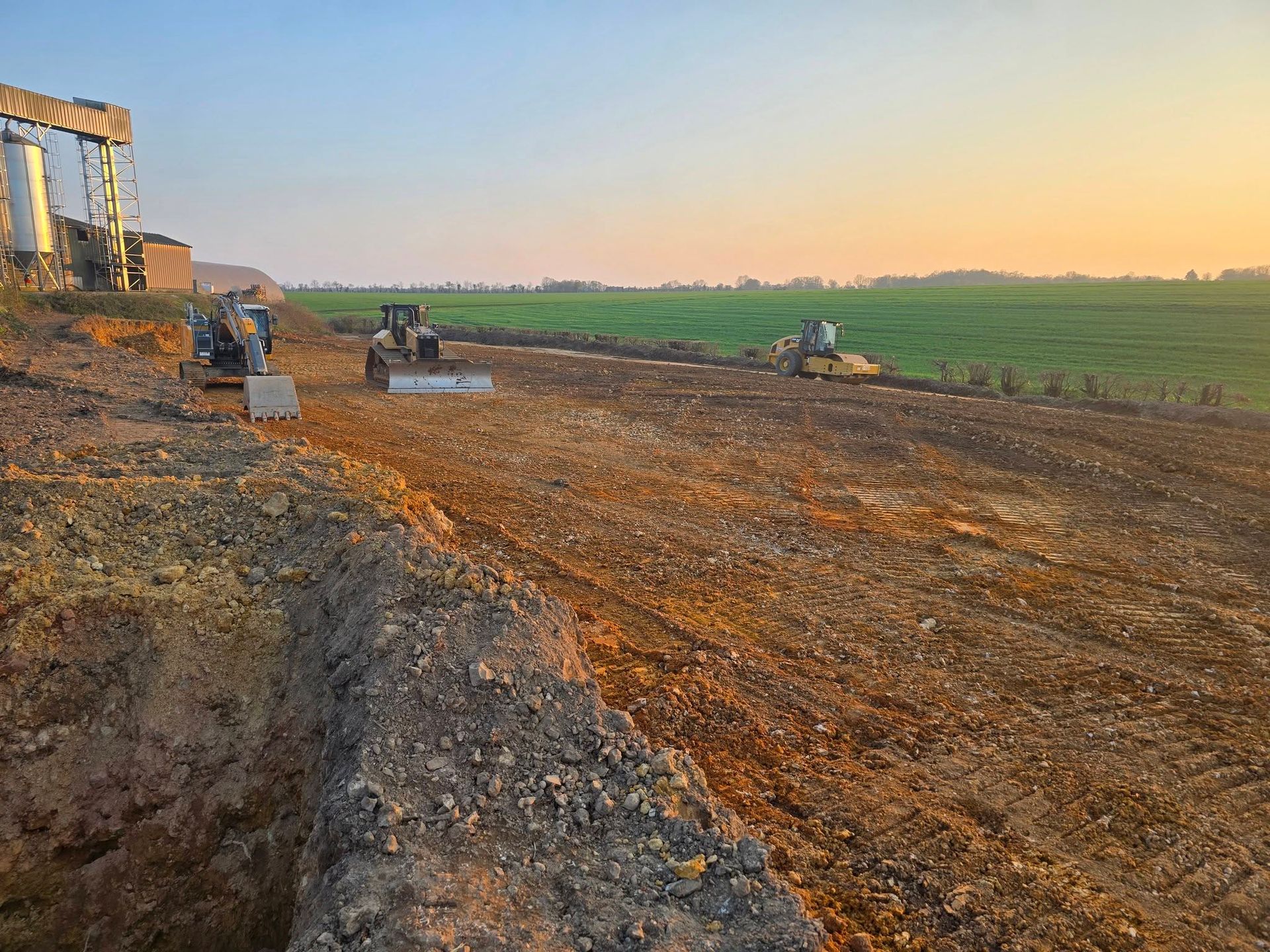 Trois engins de chantier nivellent la terre sur un site situé près d'un silo.