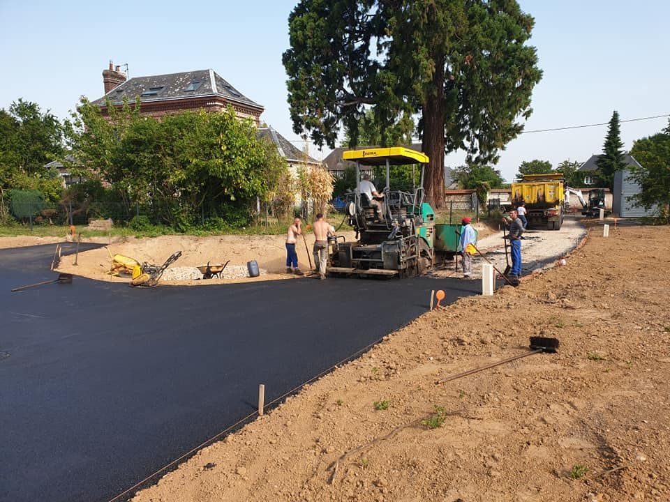 Une équipe de construction utilise une machine à paver jaune pour poser de l'asphalte noir neuf sur une route près d'une maison et d'un grand arbre.