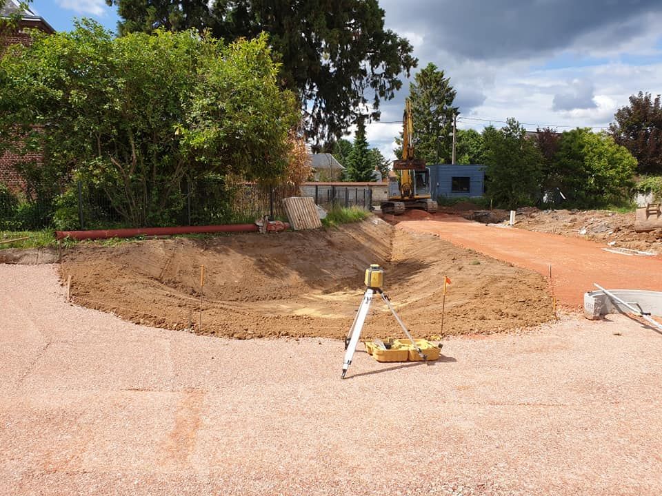 Un chantier de construction avec un outil de topographie au premier plan, un monticule de terre excavée, un chemin de gravier rouge et un conteneur bleu.