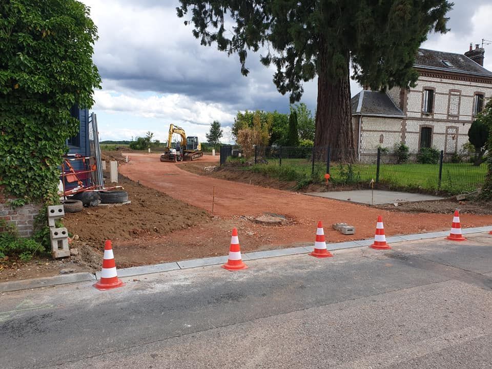 Chantier de construction avec des cônes orange bordant une allée de gravier menant à une maison, avec une excavatrice en arrière-plan.