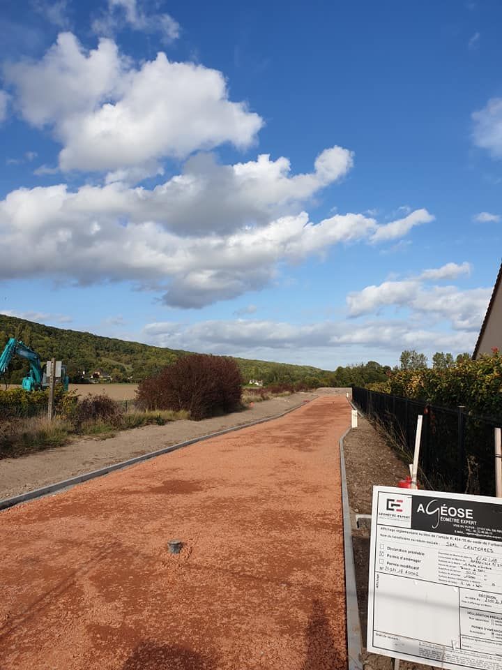 Un chemin de gravier mène à un flanc de colline boisé sous un ciel bleu parsemé de nuages, avec un panneau de chantier au premier plan.