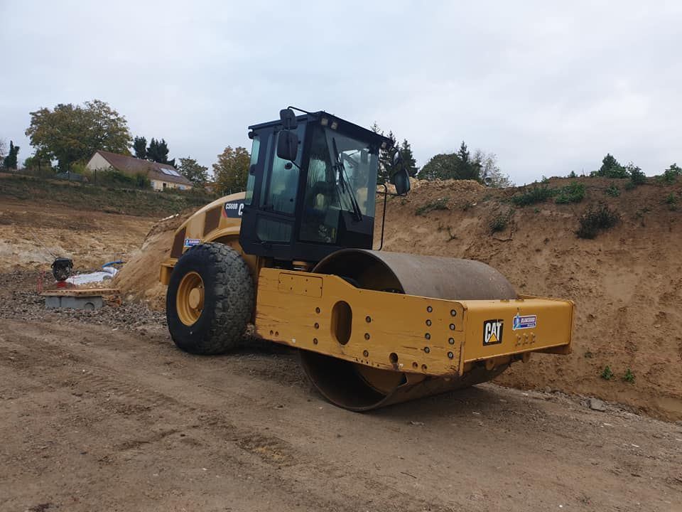 Un compacteur de sol Caterpillar jaune est stationné sur un chantier de construction, avec des monticules de terre en arrière-plan.