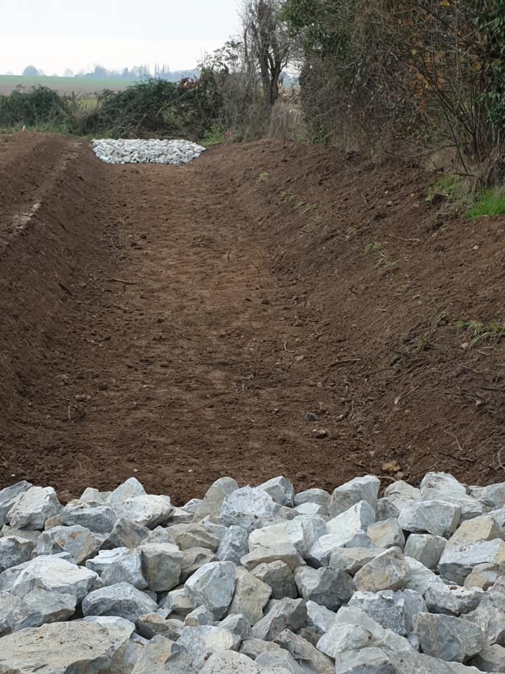 Un fossé de drainage en terre, en pente, avec des barrages de retenue rocheux en amont et en aval, dans un cadre rural.