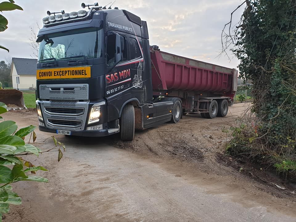 Un semi-remorque noir avec une benne basculante rouge est stationné sur un chemin de terre en zone rurale.