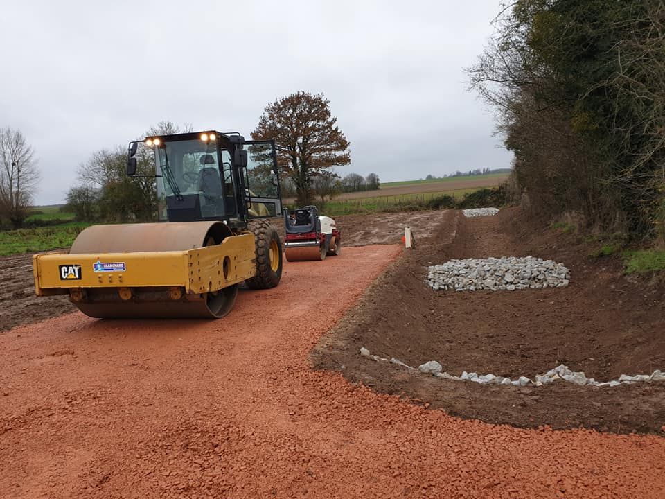 Un rouleau compresseur jaune compacte un chemin de gravier rougeâtre dans un paysage rural et ouvert.