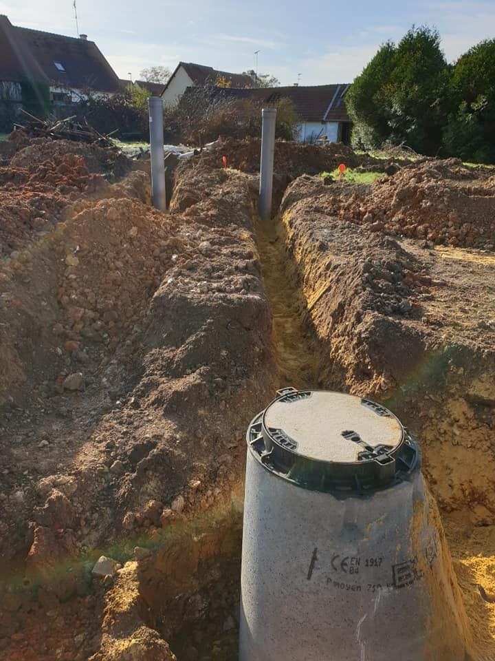 Un chantier de construction avec un regard d'égout en béton au premier plan et une tranchée profonde derrière, sous un ciel bleu.