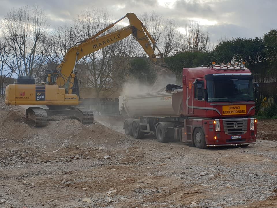 Sur un chantier de construction en plein air, une pelleteuse jaune charge de la terre dans un grand semi-remorque rouge.