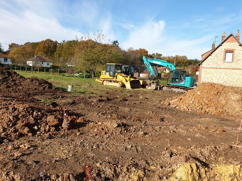 Un bulldozer jaune et une excavatrice turquoise sur un chantier de construction en terre battue, à côté d'une maison en briques.