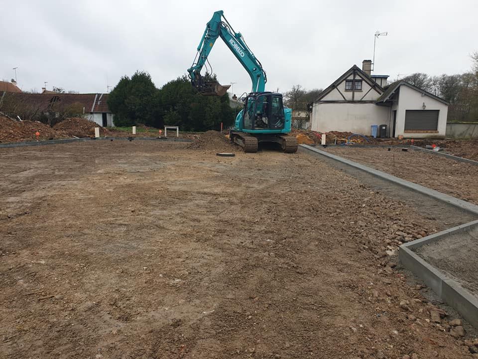 Une pelleteuse turquoise est posée sur un chantier de construction en terre battue près d'une maison, un trottoir en béton formant le périmètre.