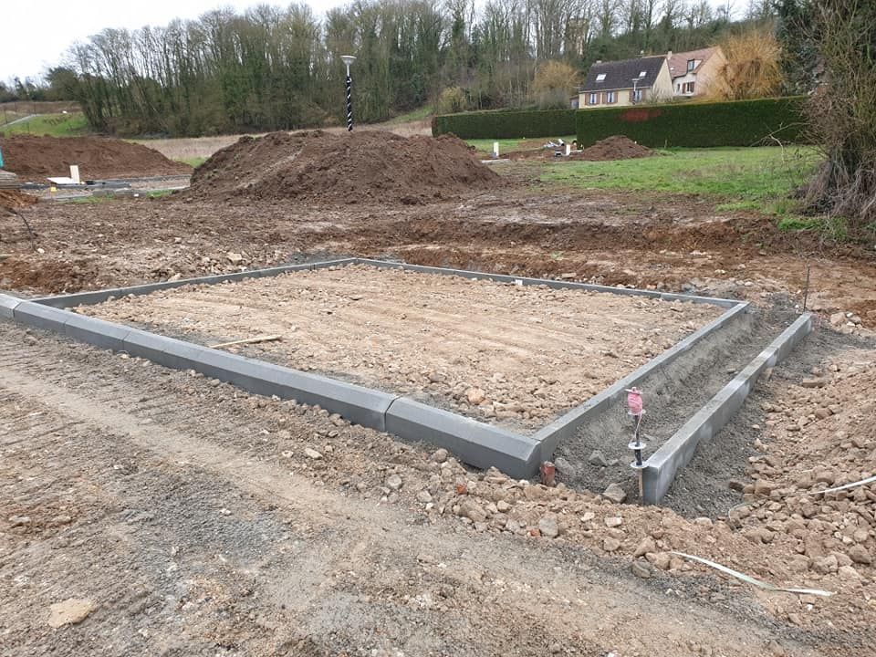 Sur un chantier de construction, une fondation rectangulaire en béton gris bordée de pierres encadre une base en terre.