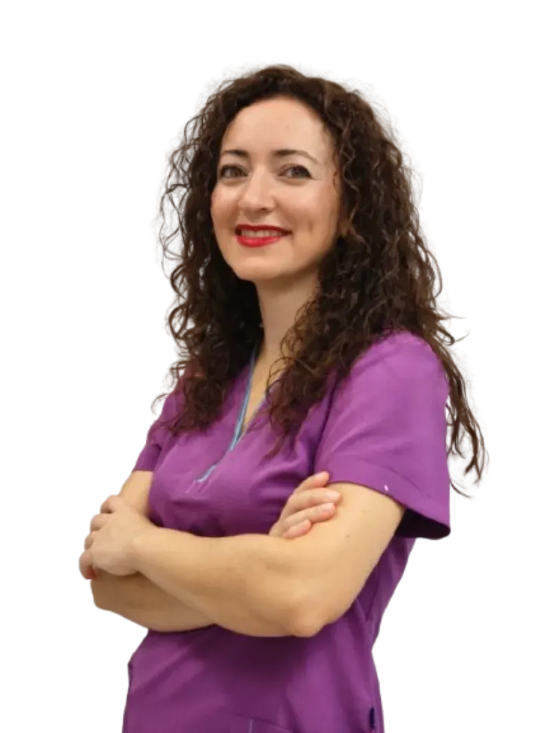 Persona sonriente con una blusa morada y los brazos cruzados, de pie sobre un fondo blanco.