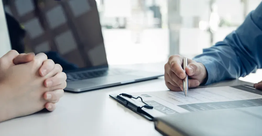 Dos personas trajeadas en un escritorio revisando documentos. Una persona firma un papel, con un mazo.