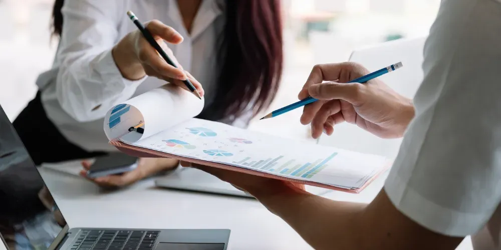 Dos personas revisando documentos, una sosteniendo un lápiz, la otra señalando con un bolígrafo, una computadora portátil.