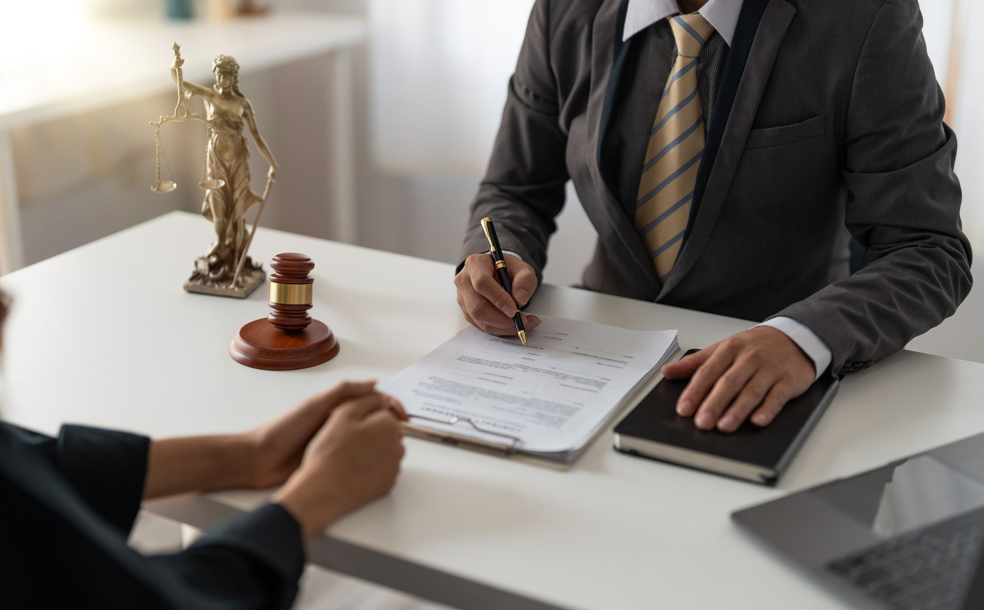Abogado escribiendo en documentos mientras consulta con un cliente, estatua de justicia y mazo sobre el escritorio.