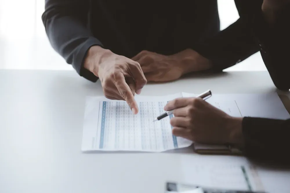 Manos apuntando a un documento sobre una mesa blanca; una mano sostiene un bolígrafo.