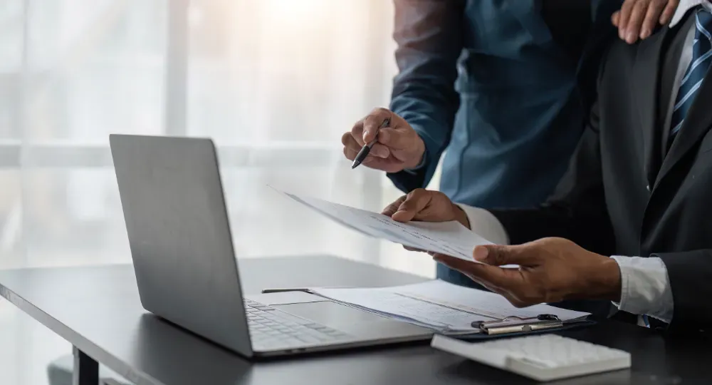 Dos personas de traje revisando documentos cerca de una computadora portátil y una calculadora en una oficina.