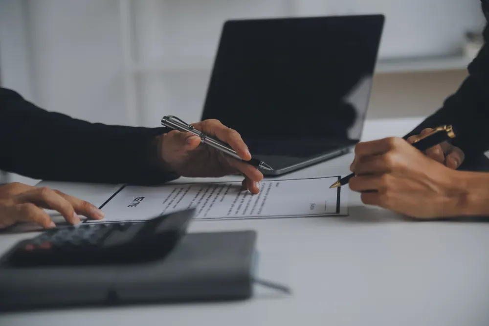 Dos personas firman un documento en un escritorio con una computadora portátil, una calculadora y un cuaderno.