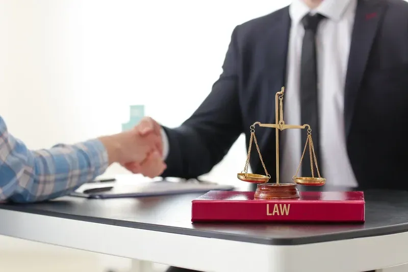 Un abogado y un cliente se dan la mano en un escritorio; en primer plano hay una balanza de la justicia.