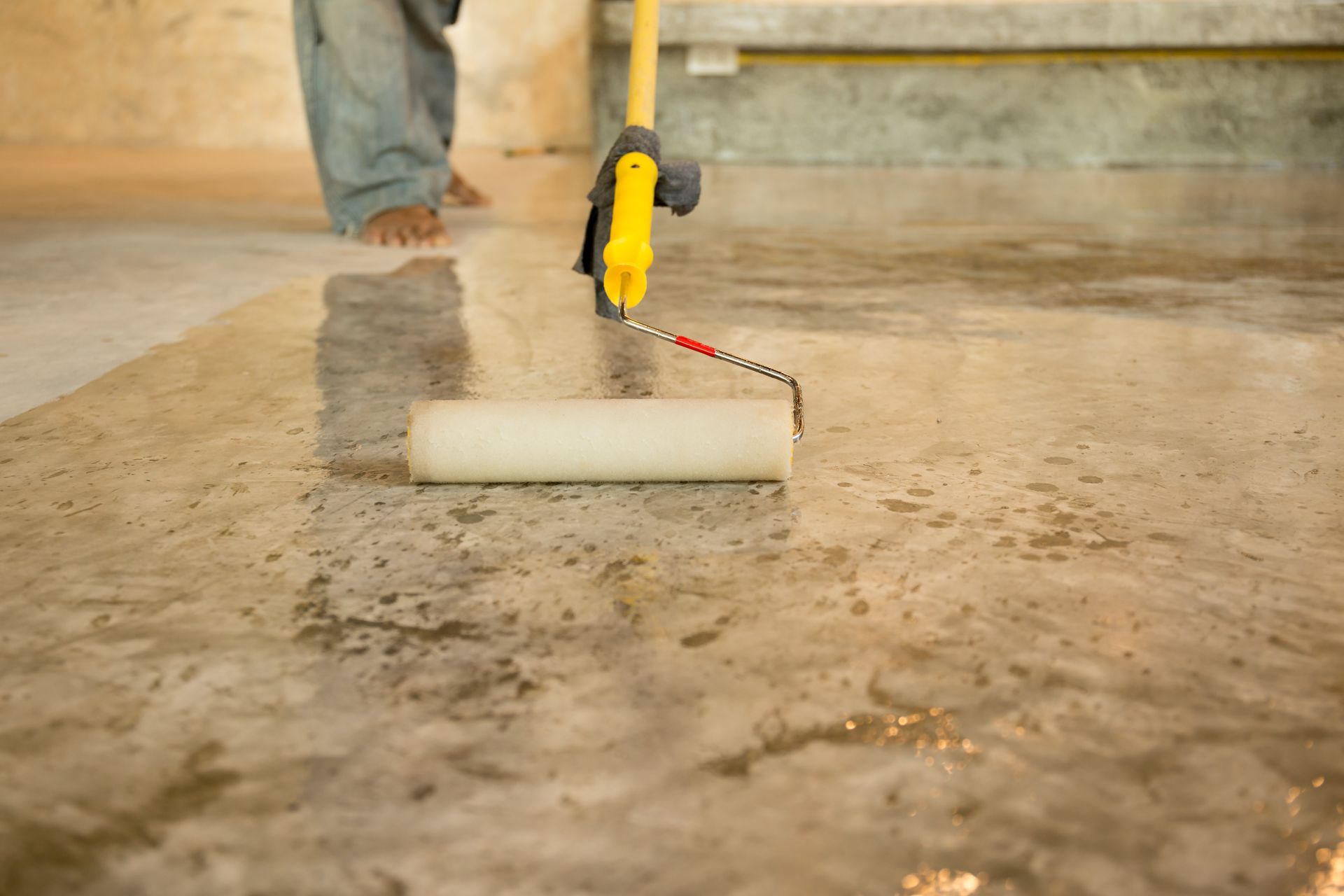Une personne utilise un rouleau à peinture à manche jaune pour appliquer un revêtement sur un sol en béton humide.